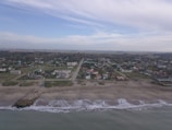 An aerial view of a coastal town with a sandy beach in the foreground. Waves gently lap the shore. Residential buildings and streets are visible, set against a backdrop of expansive greenery and a partly cloudy sky.
