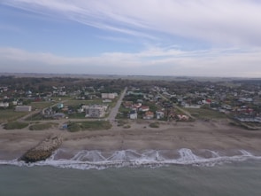 An aerial view of a coastal town with a sandy beach in the foreground. Waves gently lap the shore. Residential buildings and streets are visible, set against a backdrop of expansive greenery and a partly cloudy sky.