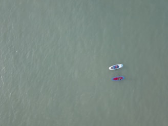 Two paddleboards float on a vast, calm expanse of water. Each board is occupied by a person, and the boards are tethered together. The water's surface is smooth, and its color is a muted green, creating a serene atmosphere.