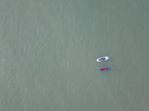Two paddleboards float on a vast, calm expanse of water. Each board is occupied by a person, and the boards are tethered together. The water's surface is smooth, and its color is a muted green, creating a serene atmosphere.