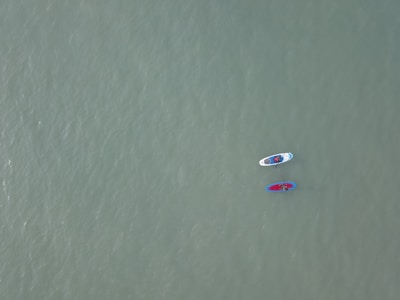 Two paddleboards float on a vast, calm expanse of water. Each board is occupied by a person, and the boards are tethered together. The water's surface is smooth, and its color is a muted green, creating a serene atmosphere.
