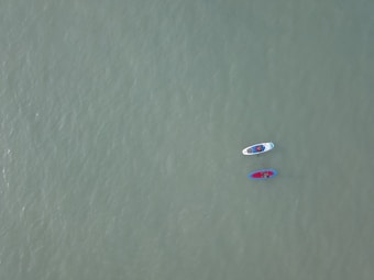 Two paddleboards float on a vast, calm expanse of water. Each board is occupied by a person, and the boards are tethered together. The water's surface is smooth, and its color is a muted green, creating a serene atmosphere.
