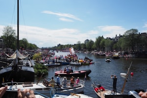 A vibrant festival scene on a canal with numerous boats carrying people decorated in colorful attire. The waterway is lined with trees and buildings on either side, and a large crowd gathers to watch the festivities. The sky is mostly clear with a few clouds, suggesting a pleasant day.