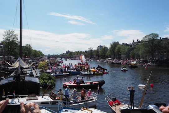A vibrant festival scene on a canal with numerous boats carrying people decorated in colorful attire. The waterway is lined with trees and buildings on either side, and a large crowd gathers to watch the festivities. The sky is mostly clear with a few clouds, suggesting a pleasant day.