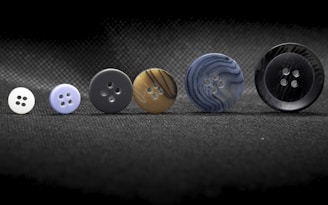A series of six buttons of varying sizes arranged in a line across a textured black surface. Each button differs in color and pattern, ranging from plain white to a marbled dark design.