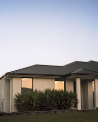 A modern house with a light-colored exterior and a dark gray, angled roof is surrounded by well-maintained greenery. Reflective windows show the orange glow of the setting sun, contrasting with the clear blue sky above.