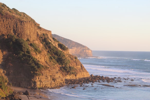 The image features a coastal landscape with tall, rocky cliffs covered in patches of green vegetation overlooking the ocean. Gentle waves roll onto the sandy shore, and the sun casts a warm, golden glow over the scene, highlighting the textures of the cliffs.