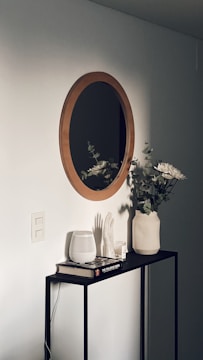 A minimalist interior scene featuring a wooden-framed round mirror mounted on a light-colored wall above a sleek, narrow console table. On the table are various decorative items, including a white vase with eucalyptus branches and flowers, a white ribbed ceramic container, a book titled 'The Polaroid Book', and a sculptural hand. The setting is accentuated by soft, natural lighting giving a tranquil ambiance.
