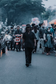 A city street scene with a man walking towards the camera, surrounded by people in casual attire. Motorcycles and cars are visible, creating a busy urban atmosphere. The crowd consists of individuals wearing jackets, masks, and some holding branded shopping bags. Trees in the background add a touch of nature.