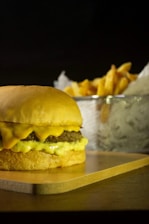 A juicy cheeseburger is placed on a wooden board, with melted cheese dripping over the beef patty. In the background, there is a metal basket filled with golden, crispy French fries.