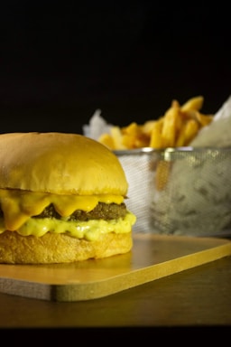 A juicy cheeseburger is placed on a wooden board, with melted cheese dripping over the beef patty. In the background, there is a metal basket filled with golden, crispy French fries.