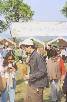 A crowd of people are gathered at an outdoor event under sunny weather. There are tents set up with a large sign overhead featuring text and illustrations. Some individuals are holding or looking at flowers, and others appear to be engaged in conversations or exploring the area. Trees and hills are visible in the background.