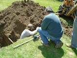 Two people are working at a dig site in a grassy area. One person is in a trench, while the other is kneeling on the ground beside it, observing or assisting. There's a backhoe visible in the background, indicating the use of heavy machinery in the digging process. Various tools are scattered around them, including a shovel.