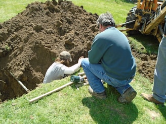 Two people are working at a dig site in a grassy area. One person is in a trench, while the other is kneeling on the ground beside it, observing or assisting. There's a backhoe visible in the background, indicating the use of heavy machinery in the digging process. Various tools are scattered around them, including a shovel.