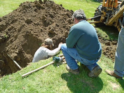 Two people are working at a dig site in a grassy area. One person is in a trench, while the other is kneeling on the ground beside it, observing or assisting. There's a backhoe visible in the background, indicating the use of heavy machinery in the digging process. Various tools are scattered around them, including a shovel.