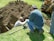 Two people are working at a dig site in a grassy area. One person is in a trench, while the other is kneeling on the ground beside it, observing or assisting. There's a backhoe visible in the background, indicating the use of heavy machinery in the digging process. Various tools are scattered around them, including a shovel.