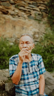 An elderly person with short gray hair is sitting outdoors against a backdrop of natural stones and greenery. They are wearing a blue plaid shirt and holding a cigarette in their hand.