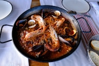 A large black paella pan filled with a seafood dish containing prawns, mussels, and clams on a bed of rice. The dish is placed on a white tablecloth, accompanied by plates, a spoon, and a basket of sliced bread.