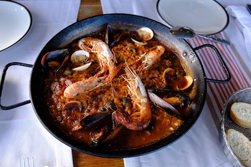 A large black paella pan filled with a seafood dish containing prawns, mussels, and clams on a bed of rice. The dish is placed on a white tablecloth, accompanied by plates, a spoon, and a basket of sliced bread.