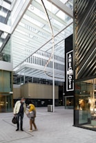 A modern architectural space with glass and metal structures, featuring an open atrium design. Two people, one wearing a face mask, are walking on a paved walkway. There is a large black sign with the word 'ANECOTE' visible on the right side. The space appears to be part of a commercial or urban area.