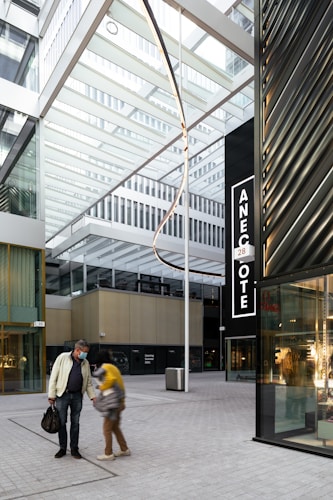 A modern architectural space with glass and metal structures, featuring an open atrium design. Two people, one wearing a face mask, are walking on a paved walkway. There is a large black sign with the word 'ANECOTE' visible on the right side. The space appears to be part of a commercial or urban area.