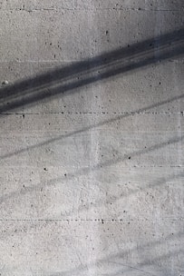 A textured concrete surface with fine grooves and scattered small holes. Diagonal shadows cast across the surface add depth and a sense of dimension.