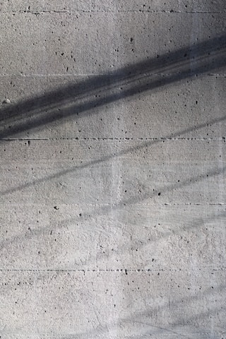 A textured concrete surface with fine grooves and scattered small holes. Diagonal shadows cast across the surface add depth and a sense of dimension.