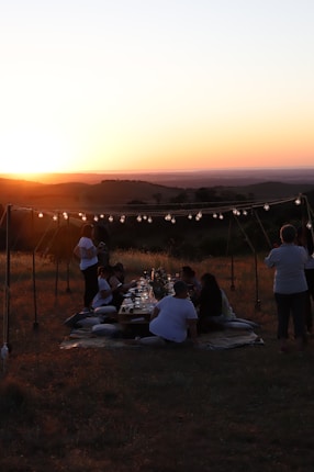 A group of people are gathered outdoors during sunset, sitting on blankets around a long table decorated with candles and string lights. The setting is an open field with a scenic view of rolling hills in the distance.