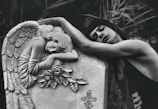 A person is leaning emotionally on a tombstone adorned with an angel sculpture and roses. The angel appears to be resting its head on its arms, conveying a sense of sorrow or mourning. The background consists of blurred foliage, adding to the serene and somber atmosphere.