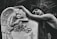 A person is leaning emotionally on a tombstone adorned with an angel sculpture and roses. The angel appears to be resting its head on its arms, conveying a sense of sorrow or mourning. The background consists of blurred foliage, adding to the serene and somber atmosphere.