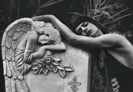 A person is leaning emotionally on a tombstone adorned with an angel sculpture and roses. The angel appears to be resting its head on its arms, conveying a sense of sorrow or mourning. The background consists of blurred foliage, adding to the serene and somber atmosphere.