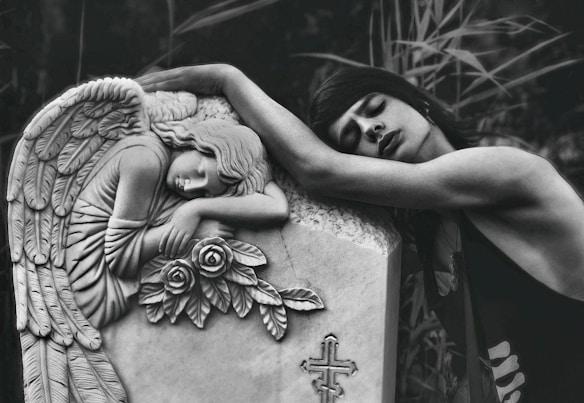 A person is leaning emotionally on a tombstone adorned with an angel sculpture and roses. The angel appears to be resting its head on its arms, conveying a sense of sorrow or mourning. The background consists of blurred foliage, adding to the serene and somber atmosphere.