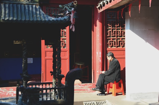 A man sits on a red stool in front of a traditional Chinese architectural structure, partially illuminated by sunlight. The architecture features intricate geometric patterns and a deep red color. Shadows and light create a serene and contemplative atmosphere.