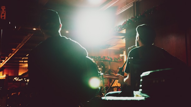 Silhouettes of two musicians, likely playing guitars, are seen performing in a dimly lit venue. Bright lights create a dramatic contrast against their outlines, and festive garlands decorate the background.