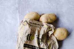 A group of potatoes partially wrapped in a brown paper bag featuring cartoon bee illustrations and eco-friendly messages, placed on a light grey surface.