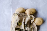 A group of potatoes partially wrapped in a brown paper bag featuring cartoon bee illustrations and eco-friendly messages, placed on a light grey surface.