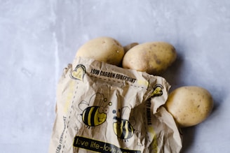 A group of potatoes partially wrapped in a brown paper bag featuring cartoon bee illustrations and eco-friendly messages, placed on a light grey surface.