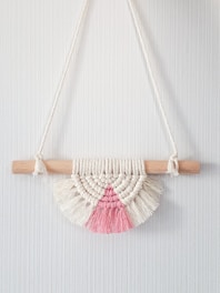 A bohemian-style macrame wall hanging featuring a semicircular design. It's crafted from cream and pink yarn, knotted around a light wooden dowel. The piece displays intricate patterns with fringe at the bottom.
