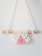A bohemian-style macrame wall hanging featuring a semicircular design. It's crafted from cream and pink yarn, knotted around a light wooden dowel. The piece displays intricate patterns with fringe at the bottom.