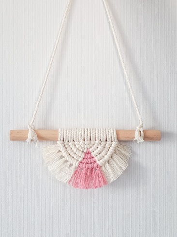A bohemian-style macrame wall hanging featuring a semicircular design. It's crafted from cream and pink yarn, knotted around a light wooden dowel. The piece displays intricate patterns with fringe at the bottom.