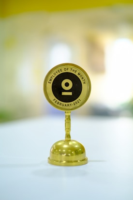 A golden trophy with a round black and gold plaque mounted on top featuring the text 'EMPLOYEE OF THE MONTH FEBRUARY-2021' placed on a light-colored surface. The background is blurred, making the trophy the focal point of the image.