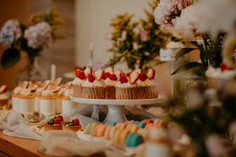 An elegantly arranged dessert table featuring an assortment of pastries and confections. A cake decorated with strawberries is prominently displayed on a white stand, surrounded by various colorful macarons, cupcakes topped with cream and fruit, and delicate floral arrangements. The table is set in a warm and inviting atmosphere with soft lighting.