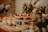 An elegantly arranged dessert table featuring an assortment of pastries and confections. A cake decorated with strawberries is prominently displayed on a white stand, surrounded by various colorful macarons, cupcakes topped with cream and fruit, and delicate floral arrangements. The table is set in a warm and inviting atmosphere with soft lighting.