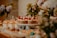 An elegantly arranged dessert table featuring an assortment of pastries and confections. A cake decorated with strawberries is prominently displayed on a white stand, surrounded by various colorful macarons, cupcakes topped with cream and fruit, and delicate floral arrangements. The table is set in a warm and inviting atmosphere with soft lighting.
