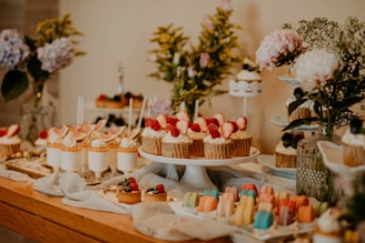 A beautifully arranged dessert table adorned with a variety of sweets, including cupcakes topped with strawberries, macarons in multiple pastel colors, and creamy desserts in small glasses. The table is decorated with floral arrangements in vases, featuring pink and purple flowers, creating an inviting and elegant setting.