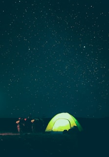 A glowing green tent is pitched under a vast, starry night sky. Beside the tent, a motorcycle is parked, adding an element of adventure to the scene. The stars twinkle brightly against the dark backdrop, creating a serene and peaceful atmosphere.