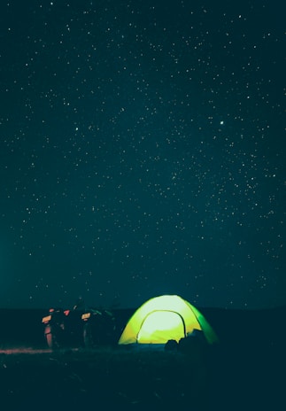 A glowing green tent is pitched under a vast, starry night sky. Beside the tent, a motorcycle is parked, adding an element of adventure to the scene. The stars twinkle brightly against the dark backdrop, creating a serene and peaceful atmosphere.