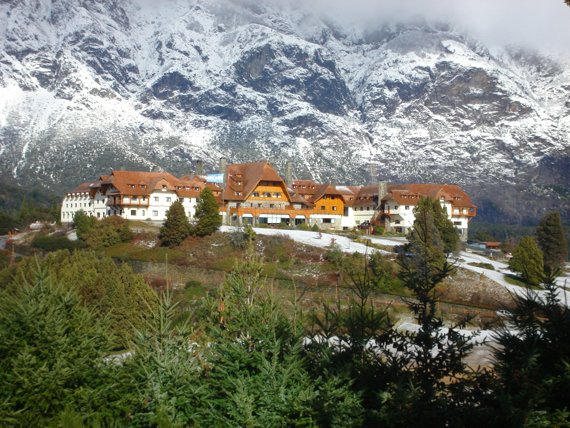 A large, rustic lodge with brown, gabled roofs is surrounded by evergreen trees. In the background, snow-covered mountains rise majestically into a cloudy sky. The foreground features a mix of greenery and patches of snow.
