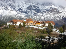 A large, rustic lodge with brown, gabled roofs is surrounded by evergreen trees. In the background, snow-covered mountains rise majestically into a cloudy sky. The foreground features a mix of greenery and patches of snow.