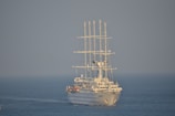 A large white sailing ship with multiple masts is navigating through calm blue waters. The ship appears modern with sleek lines and is equipped with large sails. The sky is clear, contributing to a serene maritime setting.